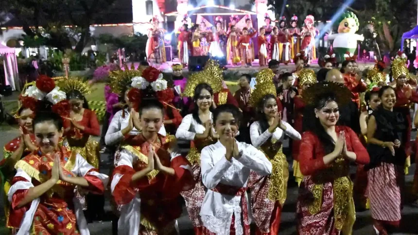 Group of people in colorful traditional costumes perform a dance at a night festival, hands pressed together in a greeting pose. A stage with performers is in the background.