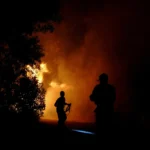 Two firefighters silhouetted at night near a large orange blaze, smoke and sparks rising behind them.