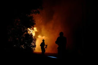 Two firefighters silhouetted at night near a large orange blaze, smoke and sparks rising behind them.