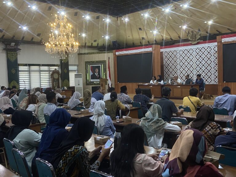 Panel of speakers at a long desk addressing an audience in a decorated conference hall with a large chandelier overhead and a portrait on the wall.