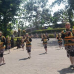 Group of dancers in green and gold traditional costumes performing a parade on a sunny day in a park or square.