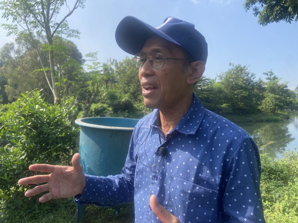 Man wearing a blue cap and glasses outdoors, gesturing with his hands, with greenery and a blue barrel behind him.