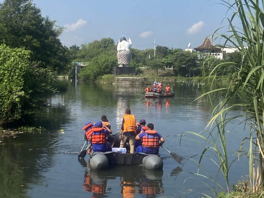 Group of people wearing orange life jackets rafting in inflatable boats on a calm river with green banks and trees nearby.