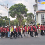 A line of students in red jackets marching down a city street, supervised by a person in a high-visibility vest, with buildings and trees in the background