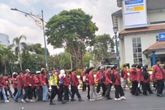A line of students in red jackets marching down a city street, supervised by a person in a high-visibility vest, with buildings and trees in the background