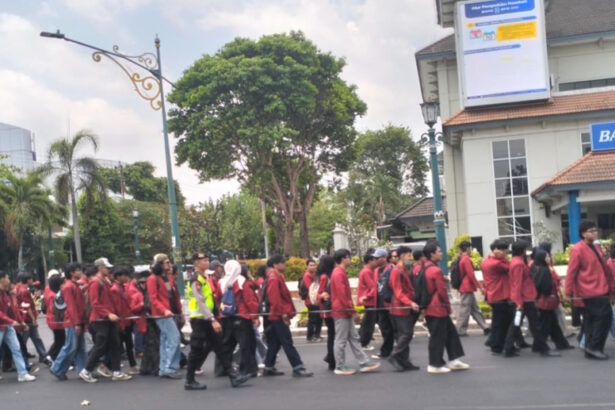 A line of students in red jackets marching down a city street, supervised by a person in a high-visibility vest, with buildings and trees in the background