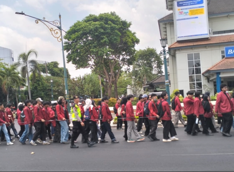 A line of students in red jackets marching down a city street, supervised by a person in a high-visibility vest, with buildings and trees in the background