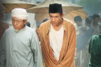 Two men in traditional clothing walk through a crowded outdoor market with umbrellas in the background.