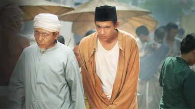 Two men in traditional clothing walk through a crowded outdoor market with umbrellas in the background.