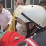 Rescue workers in a white helmet and mask attend to a person covered in red on a stretcher, as bystanders look on.