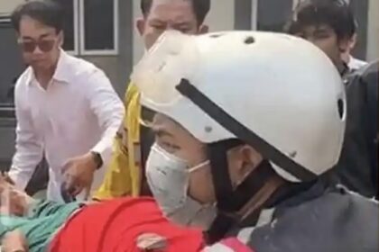 Rescue workers in a white helmet and mask attend to a person covered in red on a stretcher, as bystanders look on.