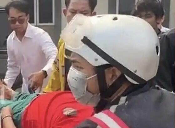 Rescue workers in a white helmet and mask attend to a person covered in red on a stretcher, as bystanders look on.