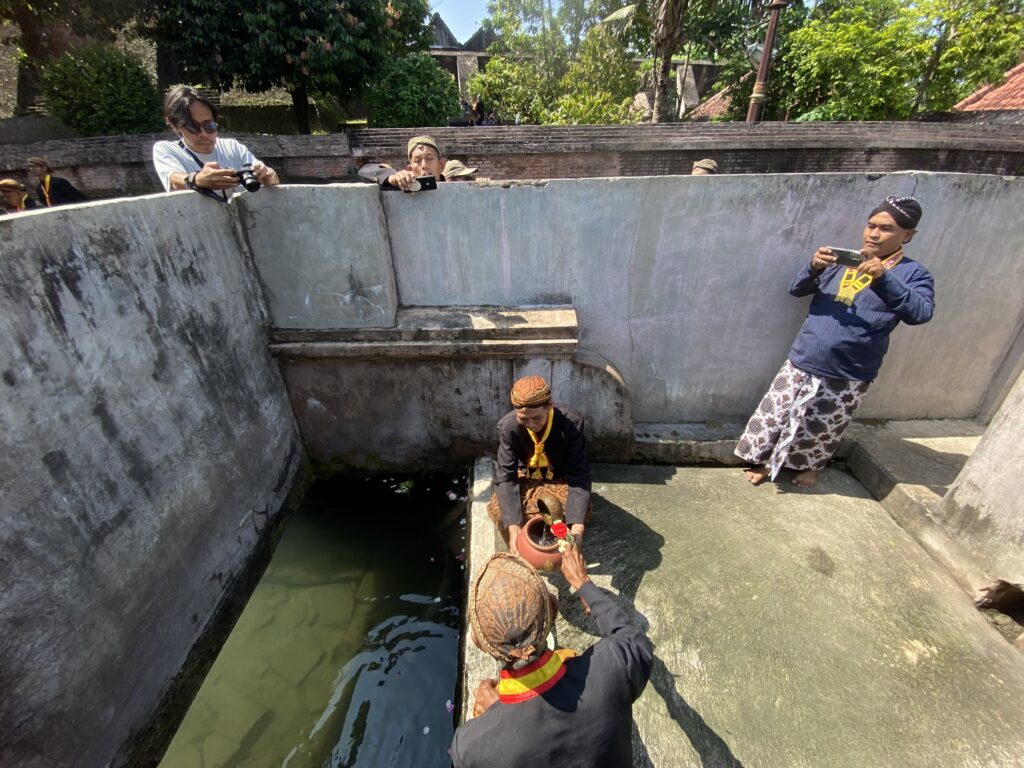 Group of people around a rectangular concrete pool performing a flower offering ritual; water below and greenery in the background.