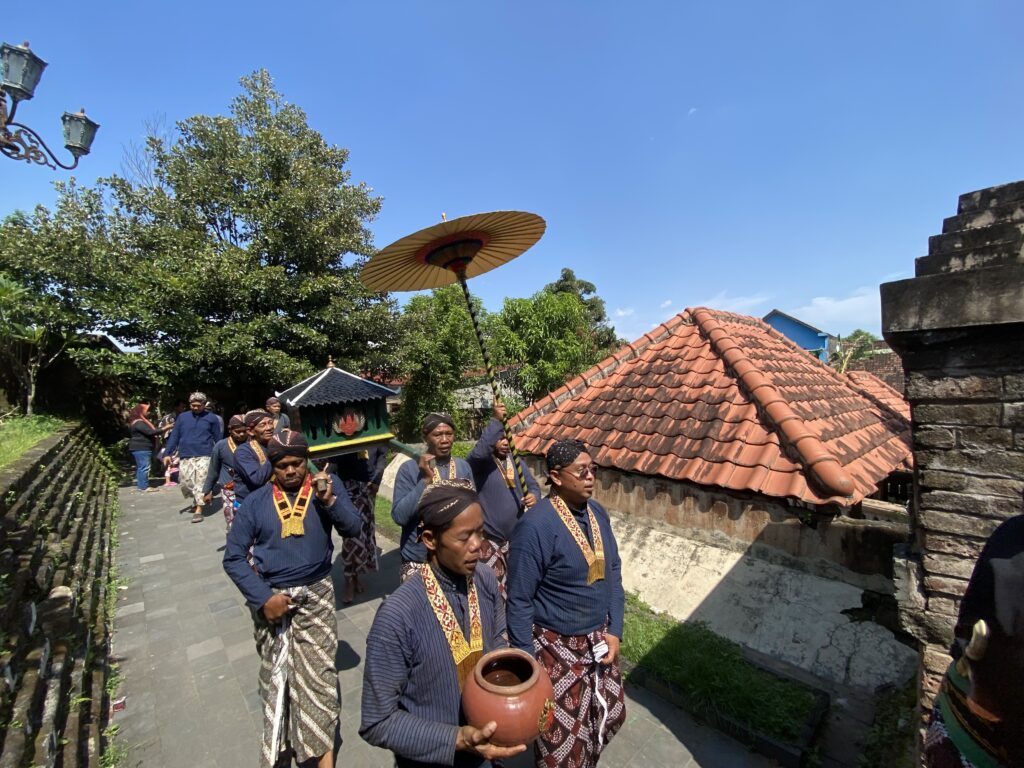 Group of people in traditional blue outfits walking a procession, carrying pottery pots and a large parasol under a bright blue sky.