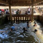 Group of children and a few adults leaning over a brick water well or canal under a wooden shelter, wading in the water below.