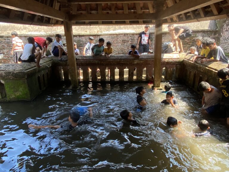 Group of children and a few adults leaning over a brick water well or canal under a wooden shelter, wading in the water below.