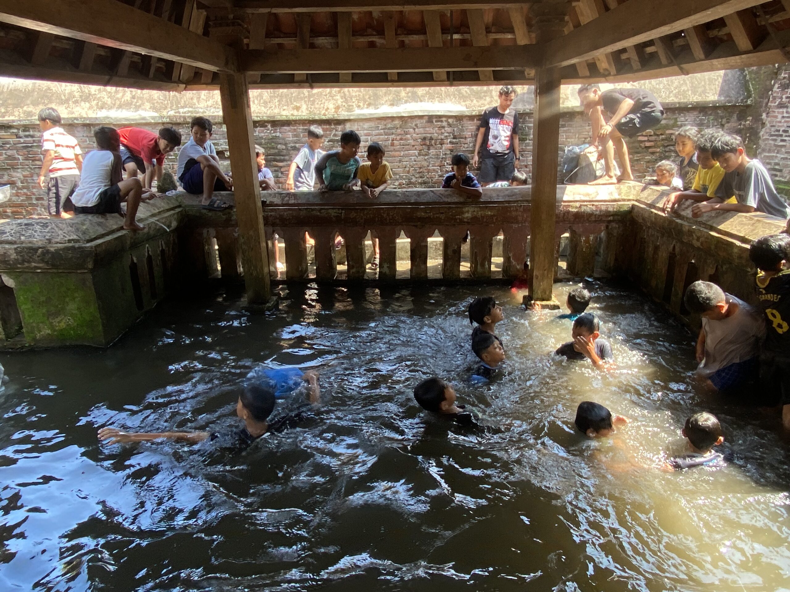 Group of children and a few adults leaning over a brick water well or canal under a wooden shelter, wading in the water below.