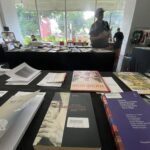 Indoor book fair or bookstore with multiple tables covered in books and magazines; shoppers browse in the bright background near large windows.
