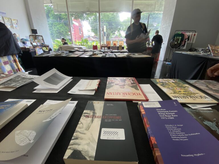 Indoor book fair or bookstore with multiple tables covered in books and magazines; shoppers browse in the bright background near large windows.