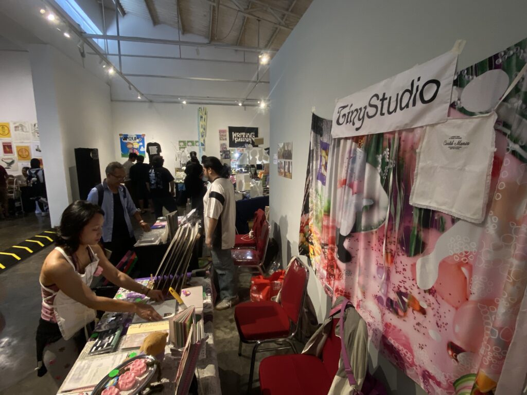 People browsing a craft fair at an indoor market with tables of supplies and colorful fabric displays behind a booth labeled GinyStudio.