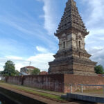 Stone multi-tiered temple tower rising from a brick enclosure beside a canal, under a bright blue sky.