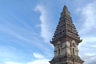 Stone multi-tiered temple tower rising from a brick enclosure beside a canal, under a bright blue sky.