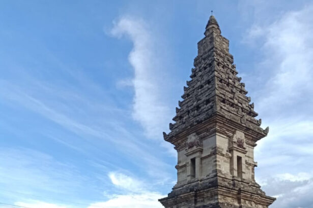 Stone multi-tiered temple tower rising from a brick enclosure beside a canal, under a bright blue sky.