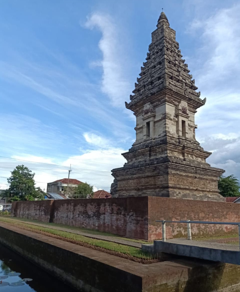 Stone multi-tiered temple tower rising from a brick enclosure beside a canal, under a bright blue sky.