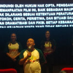 Man in green-and-white shirt speaks into a microphone on a stage, flanked by four people, with a large Indonesian text banner glowing behind them.
