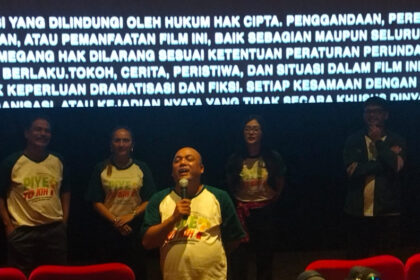 Man in green-and-white shirt speaks into a microphone on a stage, flanked by four people, with a large Indonesian text banner glowing behind them.