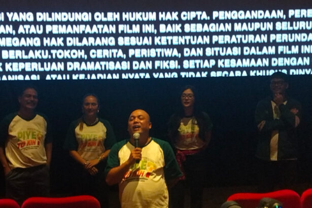 Man in green-and-white shirt speaks into a microphone on a stage, flanked by four people, with a large Indonesian text banner glowing behind them.
