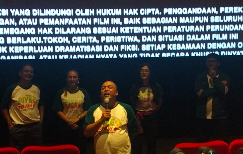 Man in green-and-white shirt speaks into a microphone on a stage, flanked by four people, with a large Indonesian text banner glowing behind them.