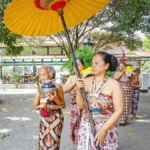 Group of Balinese women in sarongs performing a traditional ceremony under a large yellow umbrella, holding bowls.