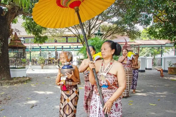 Group of Balinese women in sarongs performing a traditional ceremony under a large yellow umbrella, holding bowls.