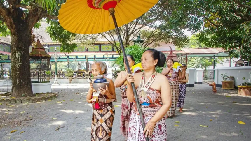 Group of Balinese women in sarongs performing a traditional ceremony under a large yellow umbrella, holding bowls.