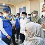 Officials and healthcare workers at a vaccination clinic; man in blue vest consults a seated woman in a hijab. Masks are worn by all present.