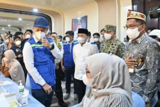 Officials and healthcare workers at a vaccination clinic; man in blue vest consults a seated woman in a hijab. Masks are worn by all present.