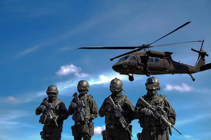 Four soldiers in black tactical gear and rifles stand in formation with a helicopter hovering in the blue sky behind them, suggesting a military operation.