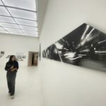 A woman in black stands in a white gallery, looking at large black-and-white panel art along the wall.