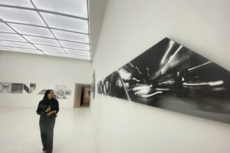 A woman in black stands in a white gallery, looking at large black-and-white panel art along the wall.