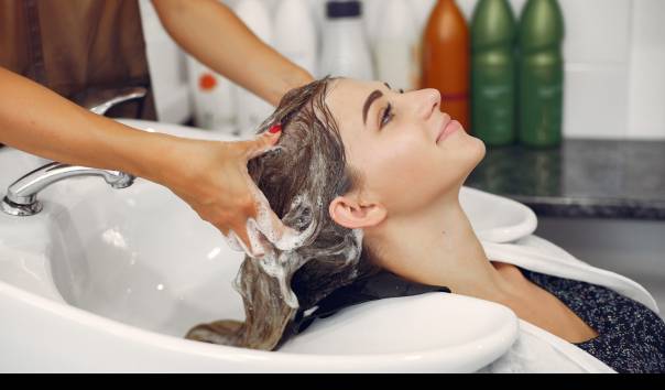 Woman reclines with head in salon sink as stylist washes her hair with shampoo bubbles present.