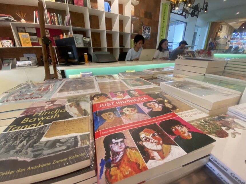 Bookstore checkout area with three people at laptops behind a display of colorful magazines to the foreground shows glossy covers; shelves in back.