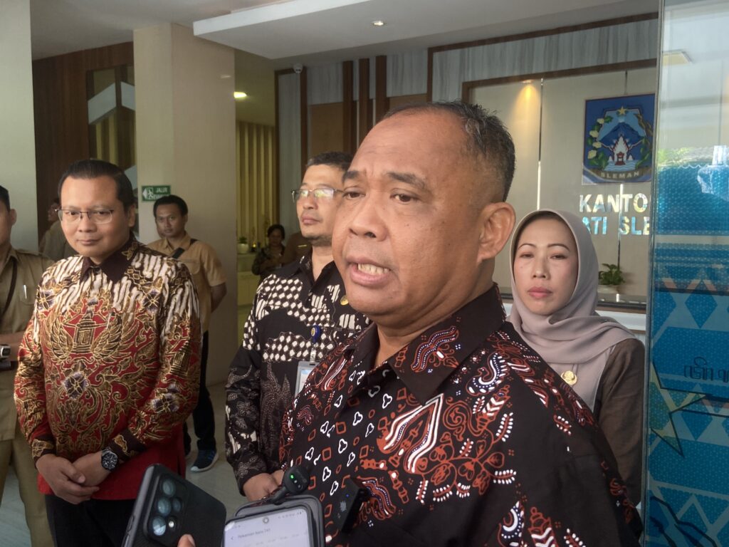 Man in a black-and-red batik shirt speaks to the camera in a group indoors; others in batik or neutral clothing stand behind in a lobby.