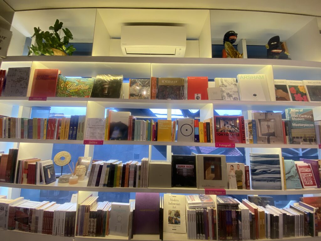 Bookstore shelves filled with colorful books and magazines, with a potted plant and decorative figures on the top shelf and an air conditioner unit above.
