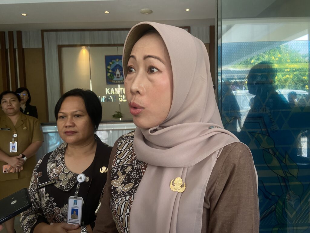 Close-up of a woman in a beige hijab and patterned top with an ID badge, in a lobby, two other staff members visible in the background.