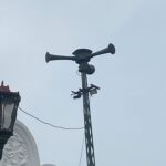 Ornate street lanterns mounted on a white building, with a tall antenna/siren apparatus on top of the structure behind them.