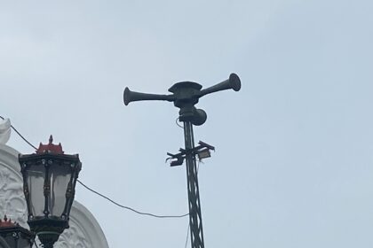 Ornate street lanterns mounted on a white building, with a tall antenna/siren apparatus on top of the structure behind them.