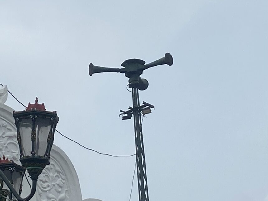 Ornate street lanterns mounted on a white building, with a tall antenna/siren apparatus on top of the structure behind them.