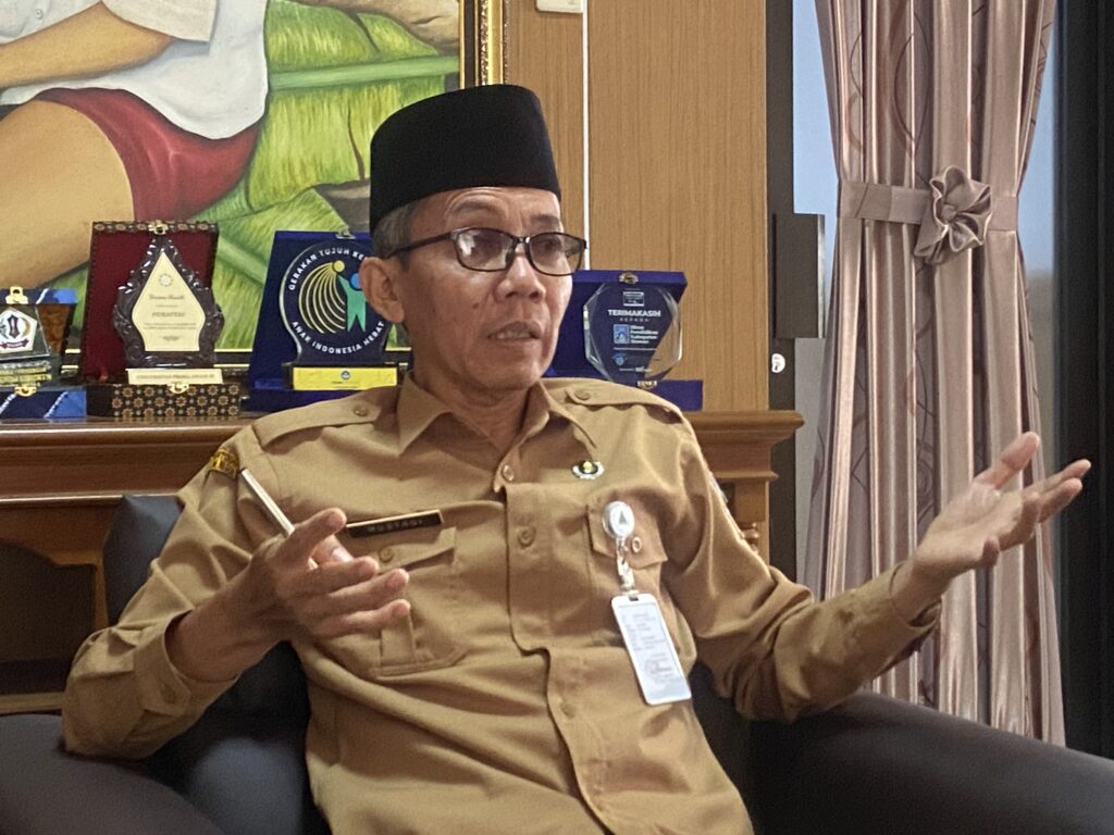 Man in tan uniform and black hat seated indoors, gesturing with hands during a discussion, awards displayed in the background.