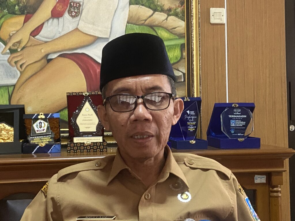 Portrait of a man in a khaki uniform and black cap, seated in front of a wall with awards and a painting in the background.